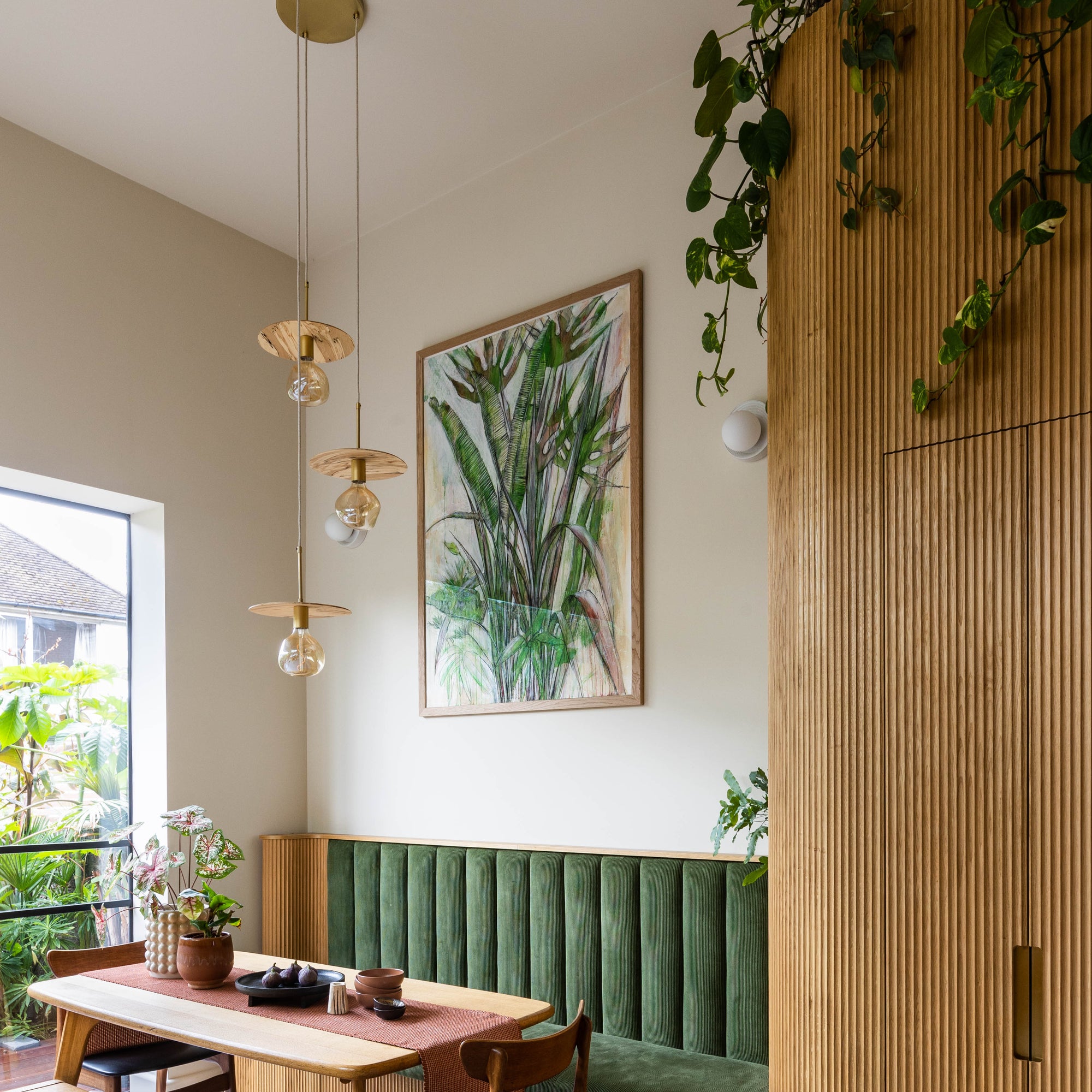 Modern dining area with wooden table, chairs, and bench against a wall with a plant painting. three wooden pendant lights