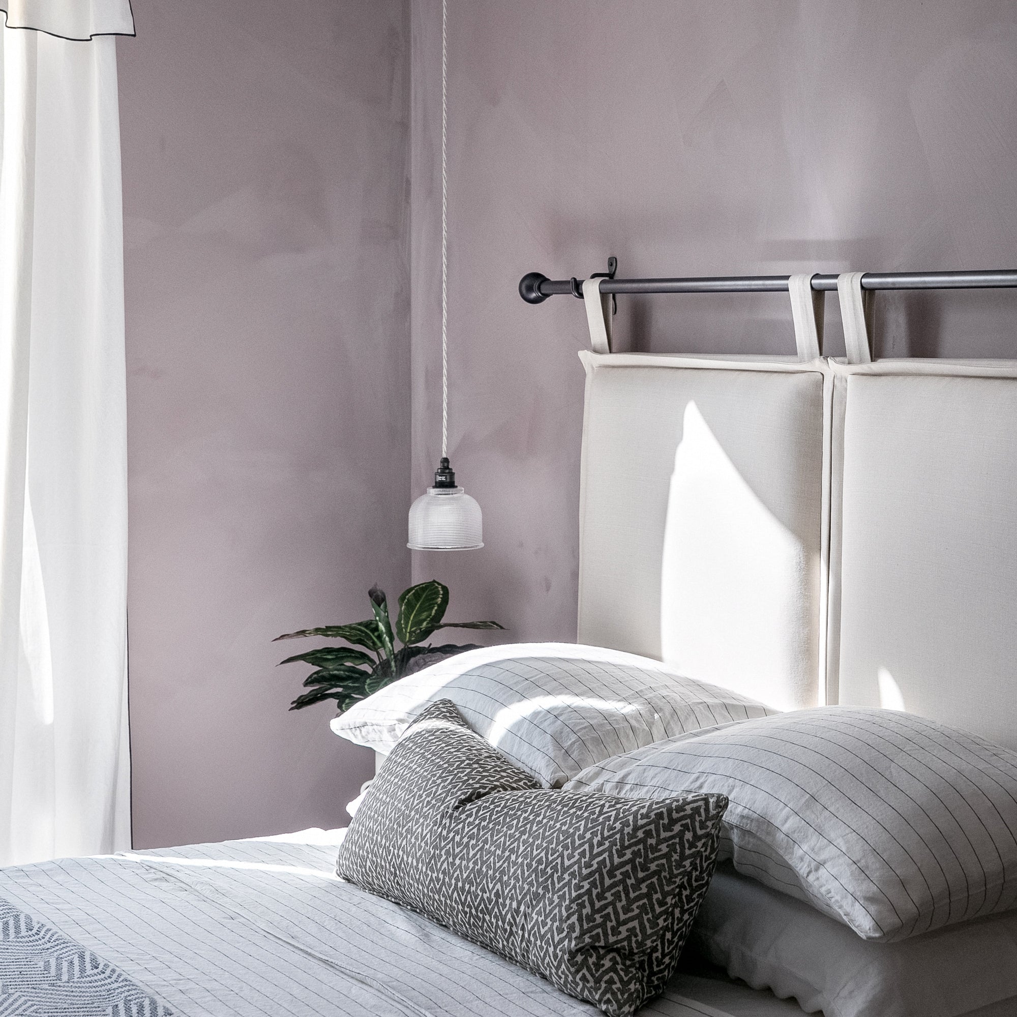 Neatly made bed with pillows and a decorative pillow against a light purple wall and hanging light