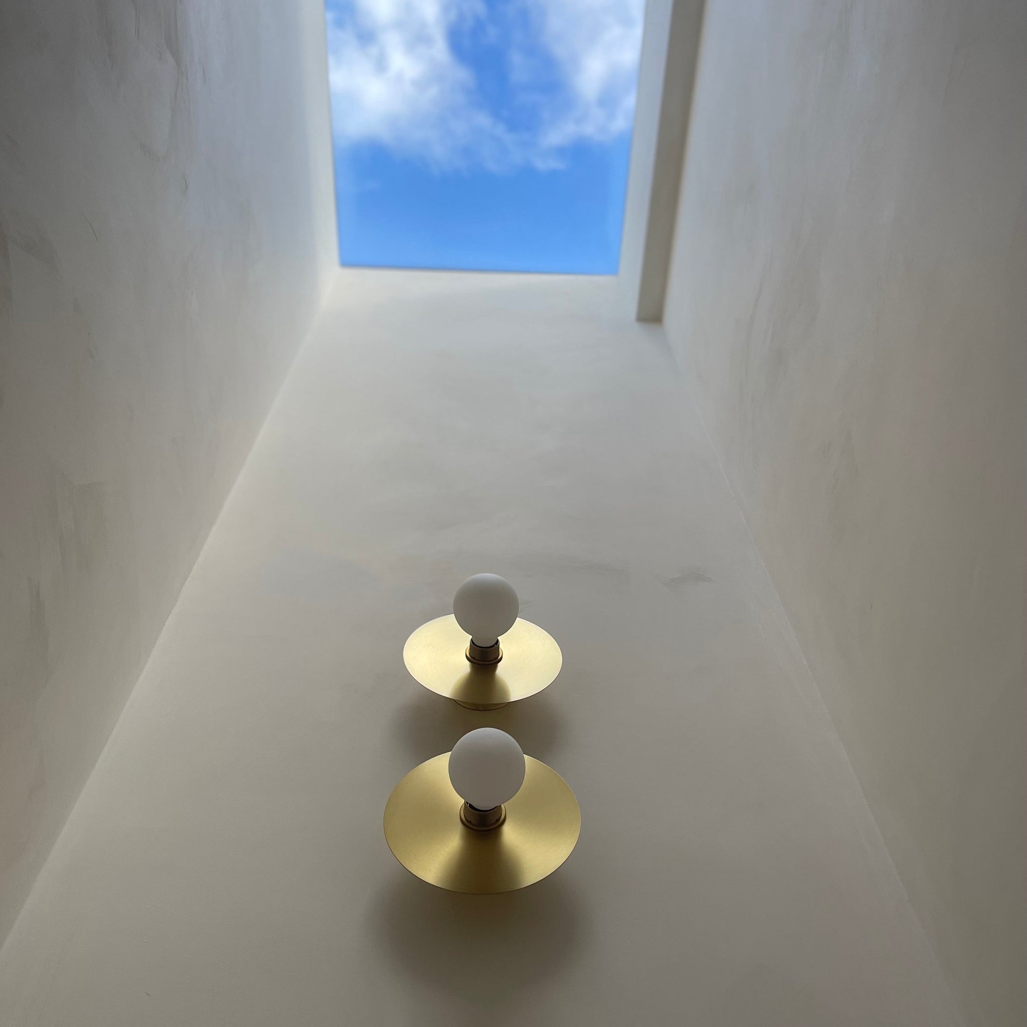 Two gold brass light fixtures with white bulbs on a white wall with a window showing blue sky.